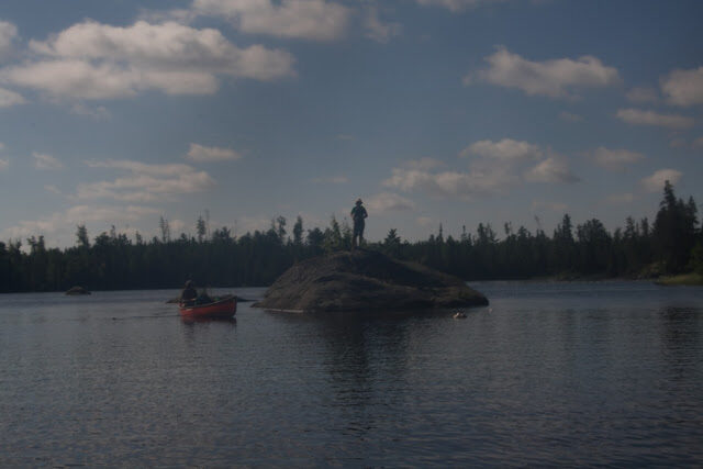 A paddler stands on a rock island beside a beached red canoe under a big dramatic sky