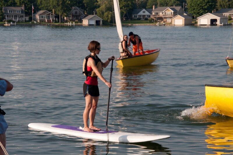 A paddleboarder and a sailboat sharing the lake in warm evening light